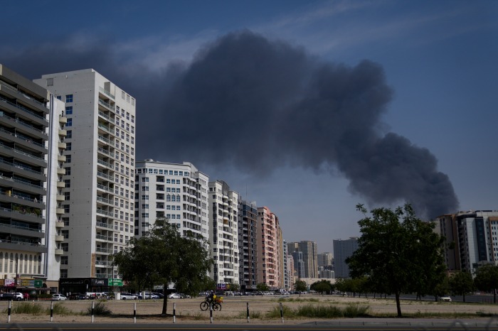 A cyclist rides past buildings in Sharjah City with a large plume of black smoke rising in the background.